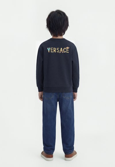 Παιδί με κοντά σκούρα μαλλιά που φοράει μαύρο φούτερ Versace, μπλε τζιν και καφέ αθλητικά παπούτσια, στέκεται με την πλάτη προς τα έξω σε λευκό φόντο.