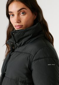 Melnā pūkainā jaka ar augstu apkakli, ar pogu aizdari un teksturētu apdari. "Pepe Jeans" logo baltā krāsā kreisajā piedurknes pusē.