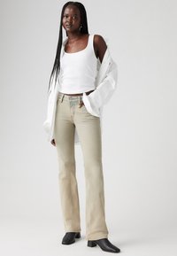 Jean flare vert clair à taille haute, associé à un top en maille blanche et une chemise blanche surdimensionnée. Bottes à talons carrés noires.