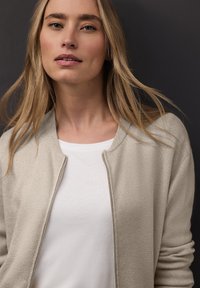 Beżowy, lekki kardigan z gładką fakturą i zapięciem na zamek, noszony na prostym białym t-shircie. Włosy są luźne i opadające.