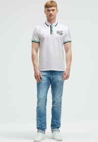 Weißes Poloshirt mit grünen und schwarzen Akzenten, ausgestattet mit einem Knopfverschluss und Logo-Detail, kombiniert mit hellblauen, verwaschenen Jeans und weißen Sneakers.