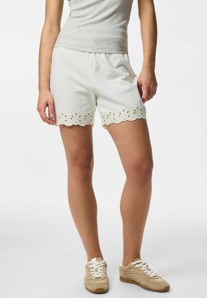 Kobieta w białych szortach z falistym, haftowanym oczkami brzegiem, szarym bezrękawnikiem i beżowymi trampkami, stojąca na jednolitym tle.