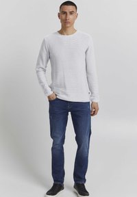 Langärmliges weißes Textshirt mit Rundhalsausschnitt, kombiniert mit dunkelblauen Jeans und schwarzen Turnschuhen. Model steht aufrecht vor einem neutralen Hintergrund.