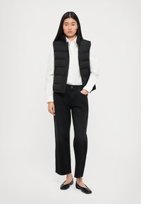 Gilet imbottito nero sopra una camicia bianca, abbinato a jeans neri a vita alta e ballerine nere. Texture morbide e vestibilità su misura.