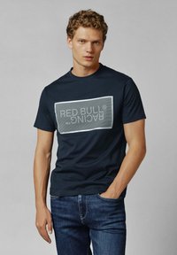 Navy katoen t-shirt met een groot rechthoekig "RED BULL RACING" logo in het grijs. Ronde hals, korte mouwen, gecombineerd met blauwe denim jeans.