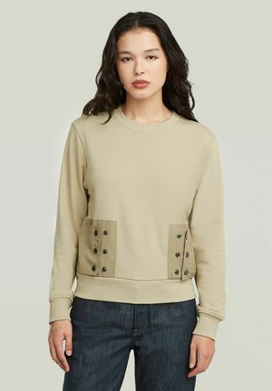 Donna con capelli lunghi e scuri indossa una felpa beige con dettagli di tasche con bottoni e jeans blu scuro su uno sfondo chiaro uniforme.