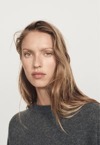 Frau mit langen blonden Haaren und grünen Augen, die einen dunkelgrauen Strickpullover trägt und direkt in die Kamera blickt, vor einem schlichten hellen Hintergrund.