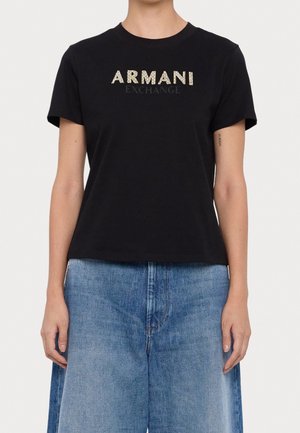 Persona que lleva una camiseta negra de Armani Exchange con texto del logo en dorado y negro, combinada con vaqueros de mezclilla azul claro.