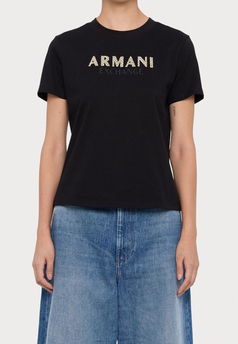 Personne portant un t-shirt noir Armani Exchange avec un logo textuel doré et noir, associé à un jean en denim bleu clair.