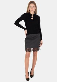 Donna che indossa un top nero a maniche lunghe con bottoni di perla e una gonna grigia con bordo in pizzo, in posa con una mano sulla vita e tacchi neri.