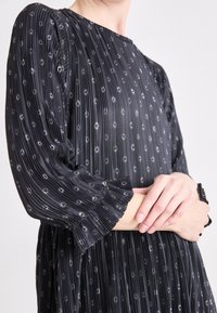 Mustmustriibuline must kleit, millel on peened täppide mustrid, kõrge kaelus ja volangidega mansetid. Sile tekstuur ja voogav siluett toovad esile disaini.