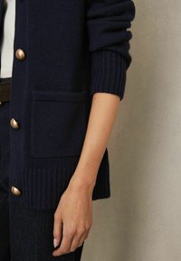 Cardigan bleumarin din material tricotat moale, cu manșete cu model în rib, un buzunar frontal și nasturi cu ton de aur pentru un finisaj elegant.