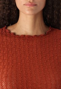 Top di colore ruggine con texture, scollatura a onde e motivo intricato, che mette in risalto il tessuto intrecciato e una sottile lucentezza.