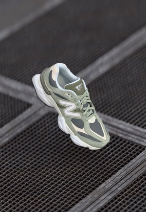 Zielono-szary but sportowy New Balance z białą podeszwą, spoczywający na czarnej metalowej podłodze z kratą i ukośnymi belkami podporowymi.