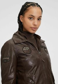 Bruine leren jas met een rits aan de voorkant, met patches en gedetailleerde stiksels. Heeft een klassieke kraag en zijzakken. Gladde textuur.