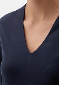 Nainen, joka pitää päällä tummansinistä V-pääntie paitaa, joka paljastaa kaulan ja ylärinnan yksinkertaista taustaa vasten.