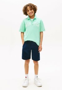 Polo verde con logo bianco, pantaloni corti blu navy, scarpe da ginnastica bianche e calzini a fantasia. La camicia presenta un colletto e una vestibilità rilassata.