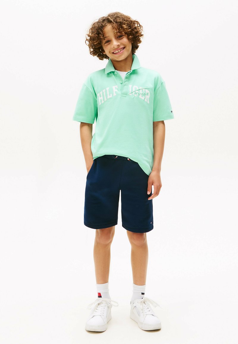 Polo verde con logo bianco, pantaloni corti blu navy, scarpe da ginnastica bianche e calzini a fantasia. La camicia presenta un colletto e una vestibilità rilassata.