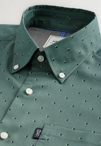 Green button-up shirt with a textured fabric, featuring small navy polka dots, a pointed collar, and a chest pocket with a logo tag.