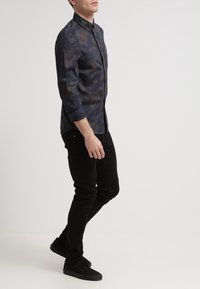 Camicia scura con motivo a fantasie e collo con bottoni, maniche lunghe, colori a contrasto, abbinata a jeans neri a taglio aderente e scarpe nere.