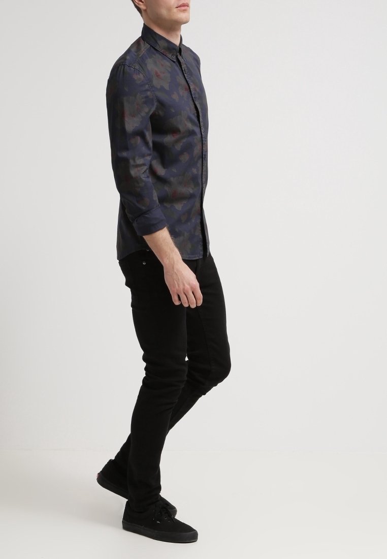 Camicia scura con motivo a fantasie e collo con bottoni, maniche lunghe, colori a contrasto, abbinata a jeans neri a taglio aderente e scarpe nere.