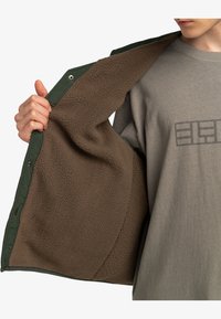 Groene doorgestikte jas met bruine fleece voering, gedeeltelijk open om de zachte textuur te laten zien. Grijze shirt eronder met een bedrukt ontwerp.
