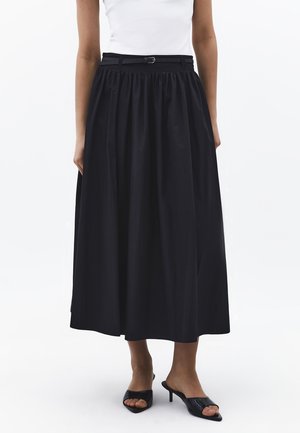 Frau trägt einen schwarzen, gerafften Midirock mit dünnem schwarzem Gürtel, weißes ärmelloses Oberteil und schwarze offene Sandalen mit Absatz, steht auf weißem Boden.