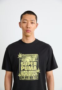 Black cotton t-shirt featuring a large yellow graphic print of a cartoon character and text reading "SUPER PUMA WORLD TOUR." Short sleeves.