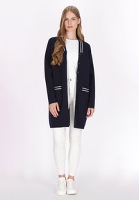 Marineblauer Cardigan mit weißen gestreiften Akzenten, mit Knöpfen an der Vorderseite und zwei Fronttaschen, kombiniert mit weißen Hosen und Schuhen.