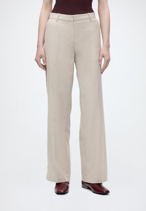 Pantalon beige à jambes larges porté avec un haut foncé et des chaussures pointues bordeaux sur une personne debout devant un fond blanc.
