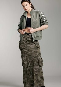 Chaqueta de chándal verde oliva con cierre de cremallera y rayas blancas, emparejada con una falda cargo de camuflaje que cuenta con bolsillos y un cinturón ancho. Top negro corto por debajo.