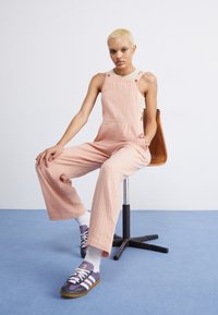 Ljust rosa bomullsbyxor med vida ben och framfickor, bärs över en beige topp. Modellen har på sig lila sneakers med vita detaljer och strumpor.