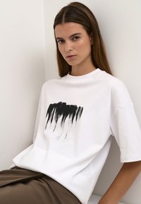 Tricou alb din bumbac, cu un design din pensulă neagră pe față, cu o croială lejeră și mâneci scurte, asociat cu o fustă maro.