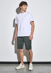 Camiseta blanca de algodón, de corte relajado, combinada con pantalones cortos verde oscuro, por encima de la rodilla. Zapatillas beiges con acentos claros completan el conjunto.