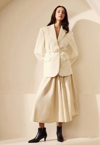 Crème blazer met een riem en oversized revers, gecombineerd met een vloeiende, knie-lange crème rok. Zwarte enkellaarsjes zorgen voor contrast.