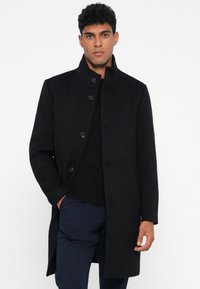 Abrigo de mezcla de lana negra con cuello alto, cierre frontal de botones y bolsillos laterales. Corte estructurado con textura suave y diseño minimalista.