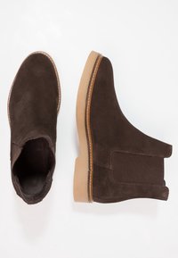 Botas de ante marrón hasta el tobillo con paneles laterales elásticos, suela de color marrón claro y costuras en contraste, con diseño de punta redonda.