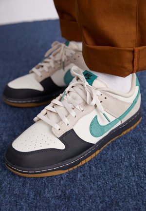 Personne portant des baskets Nike blanches, noires et bleu sarcelle avec des lacets blancs et un pantalon marron, debout sur un sol recouvert d’un tapis bleu.