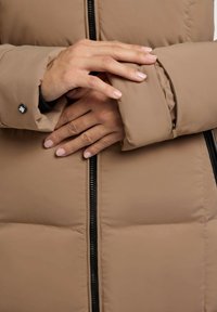 Barna puffas dzseki texturált külsővel, cipzáras záródással és gombos részletekkel az ujjakon. Töltött minta a szigetelés érdekében.