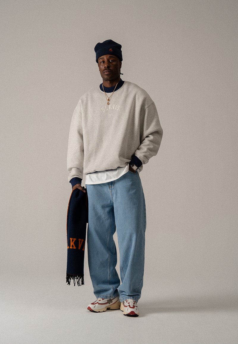 Grauer übergroßer Pullover mit gesticktem Text, hellblaue Baggy-Jeans und mehrfarbige Sneakers. Hält einen marineblauen Schal mit orangefarbener Schrift.