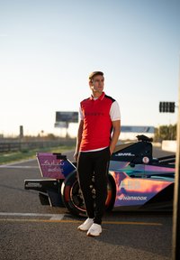 Rot-weiße Polo-Shirt mit schwarzen Akzenten, schwarze Jogginghose und weiße Schuhe, steht neben einem bunten Rennwagen in einer Außenumgebung.