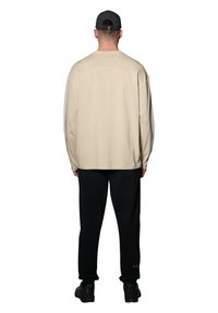 Mężczyzna w beżowej koszuli z długim rękawem, czarnych spodniach z logo przy kostce, czarnych trampkach i czarnej czapce z daszkiem, stoi tyłem do widza.