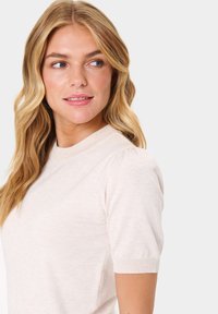 Světle béžový pletený top s krátkým rukávem, kulatým výstřihem a nabíranými detaily na rukávech. Měkká textura s přiléhavým střihem.