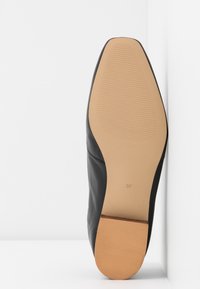 Chaussure en cuir noir avec une texture lisse et un bout arrondi, dotée d'une semelle en caoutchouc tan avec des détails nervurés et un marquage de taille.