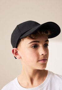 Juoda kepuraitė su lenktu snapeliu ir struktūruota karūna, pagaminta iš audinio, su maža reguliuojama juostele gale.