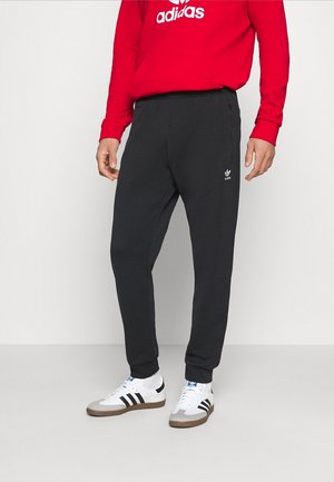 Zwarte joggingbroek met een elastische tailleband en zijzakken, voorzien van een klein wit Adidas-logo. Gecombineerd met een rode hoodie en witte sneakers.
