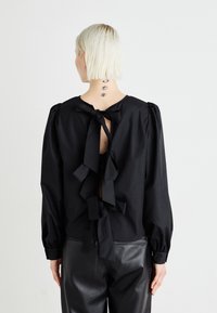 Blusa nera con maniche lunghe a sbuffo, dettagli arricciati e chiusura con lacci sul retro, realizzata in tessuto morbido, abbinata a pantaloni di pelle nera.