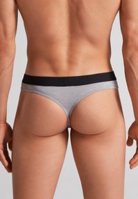 Grå bomulds-tanga med en sort elastik, der har en glat tekstur og minimalistisk design. Bagfra fremhæver beskrivelsen snittet og pasformen på tøjet.