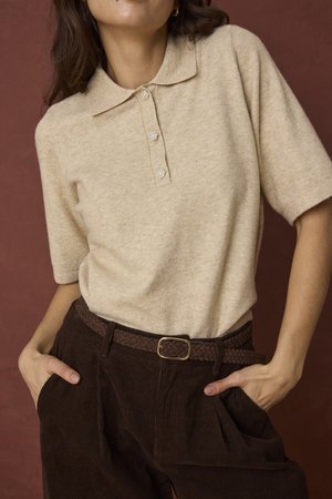 Personne portant un polo beige à manches courtes avec des boutons décoratifs et un pantalon plissé marron foncé avec une ceinture tressée, mains dans les poches.