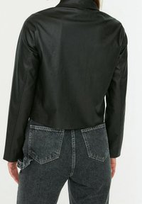 Zwarte neplederen cropped jack met een gladde textuur, lange mouwen en een eenvoudige kraag, gedragen over donkere spijkerbroeken met rafeldetails.